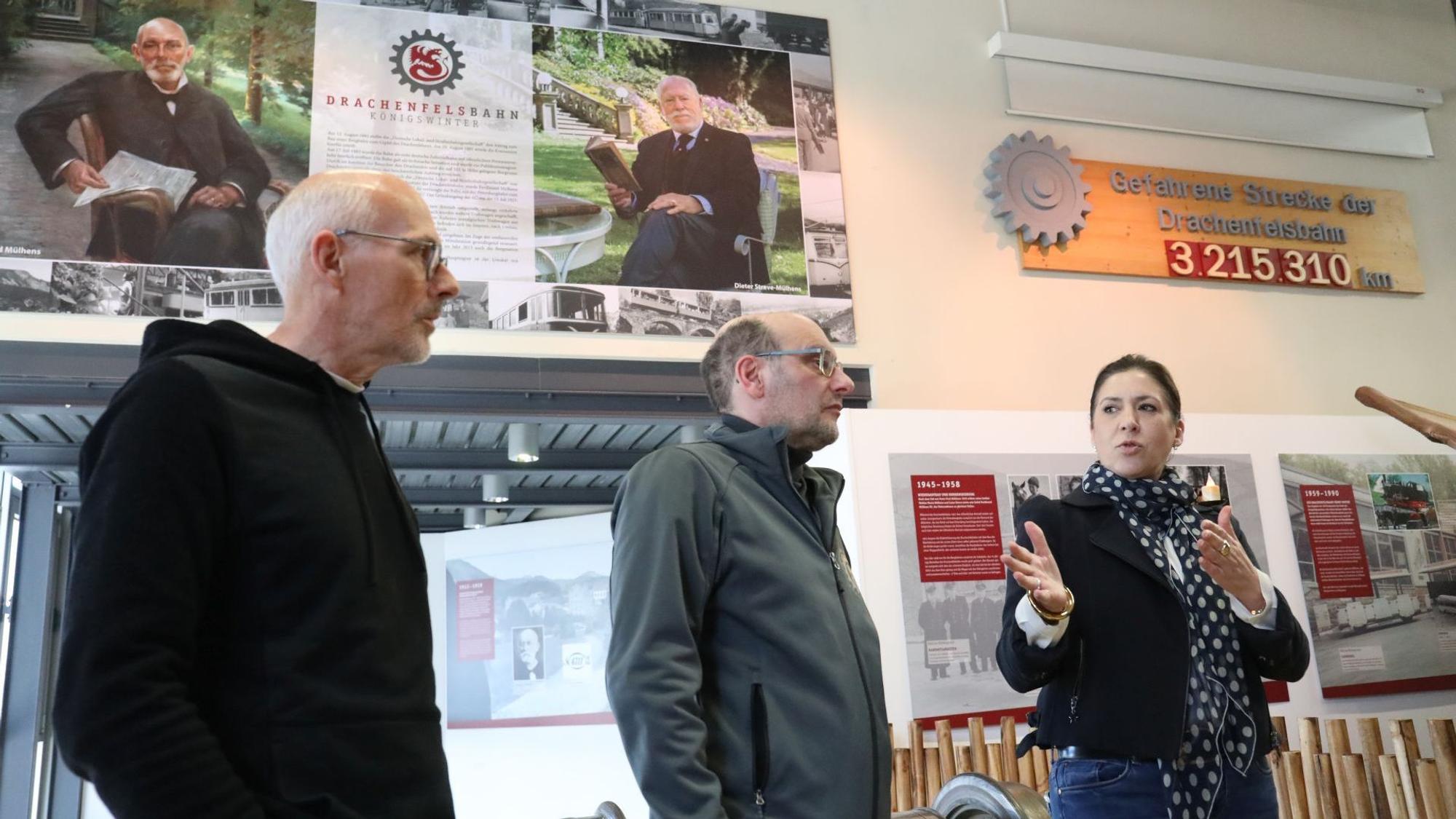 Zwei Männer und eine Frau, im Hintergrund historische Aufnahmen einer Ausstellung.