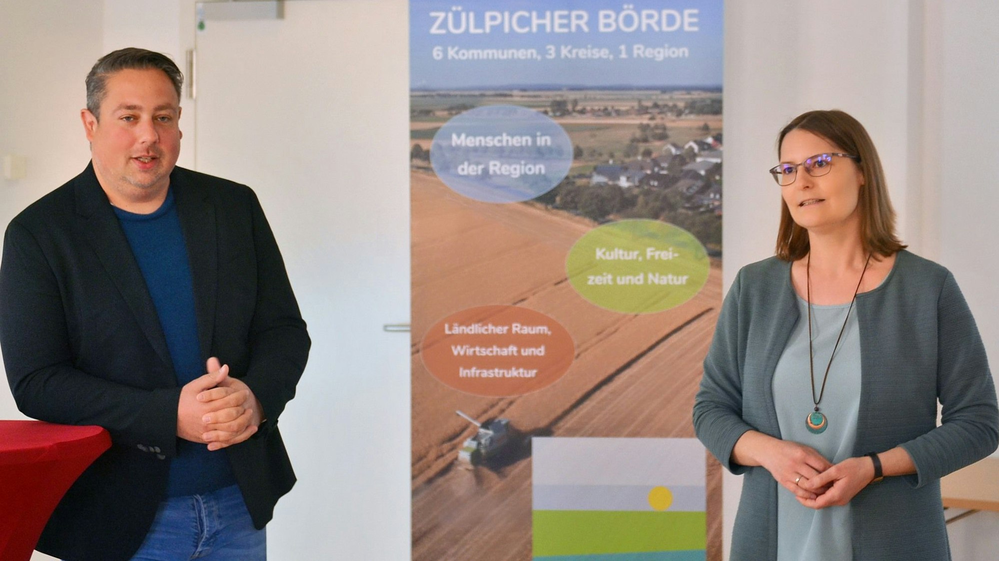 Jens Alvermann lehnt an einem Stehtisch, Silvia Bruder neben einem Roll-up-Display mit der Aufschrift „Zülpicher Börde“.