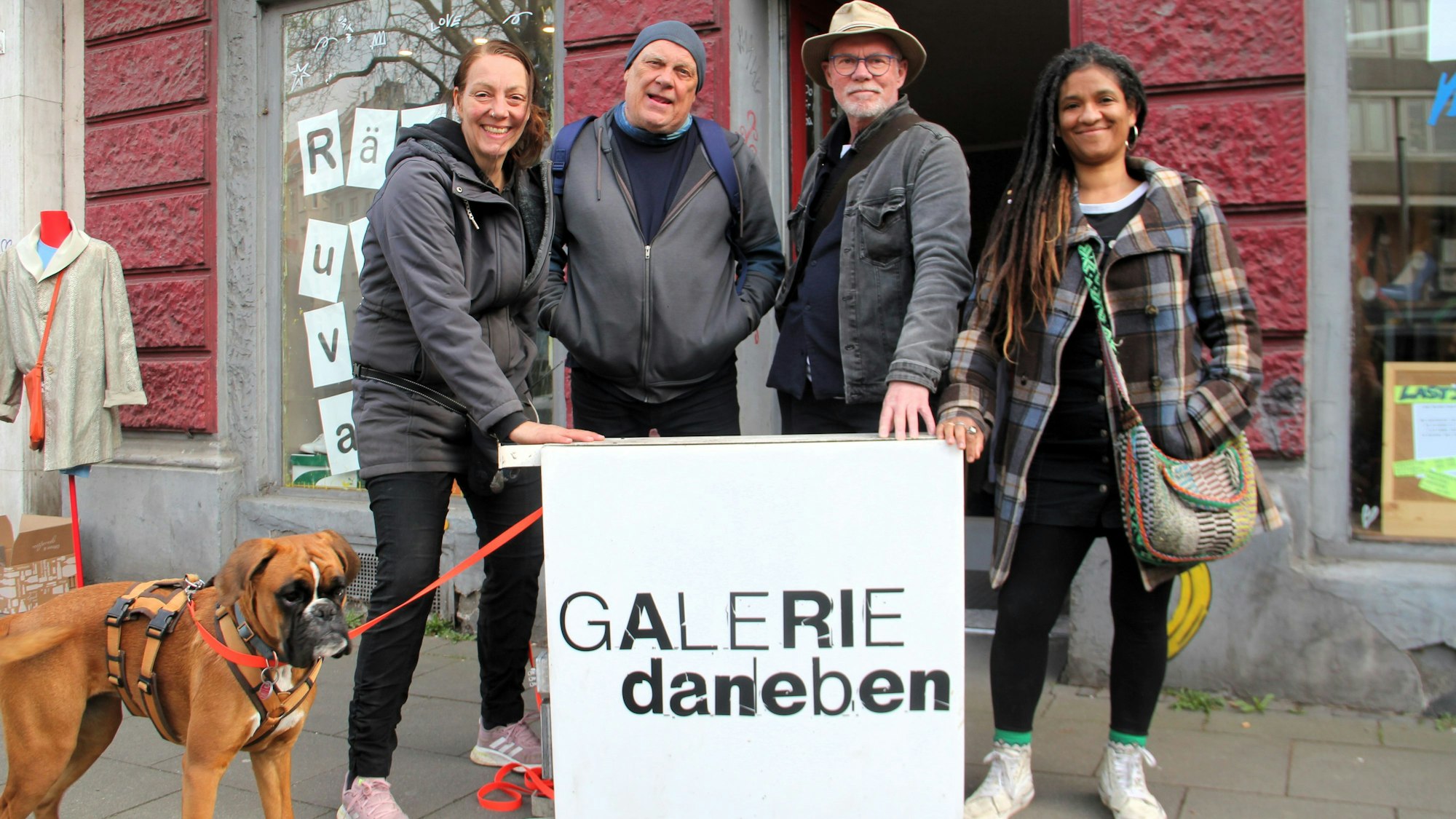 Vier Leute und ein Hund stehen auf einem Bürgersteig. Vor ihnen ist ein Schild mit dem Schriftzug Galerie Daneben.