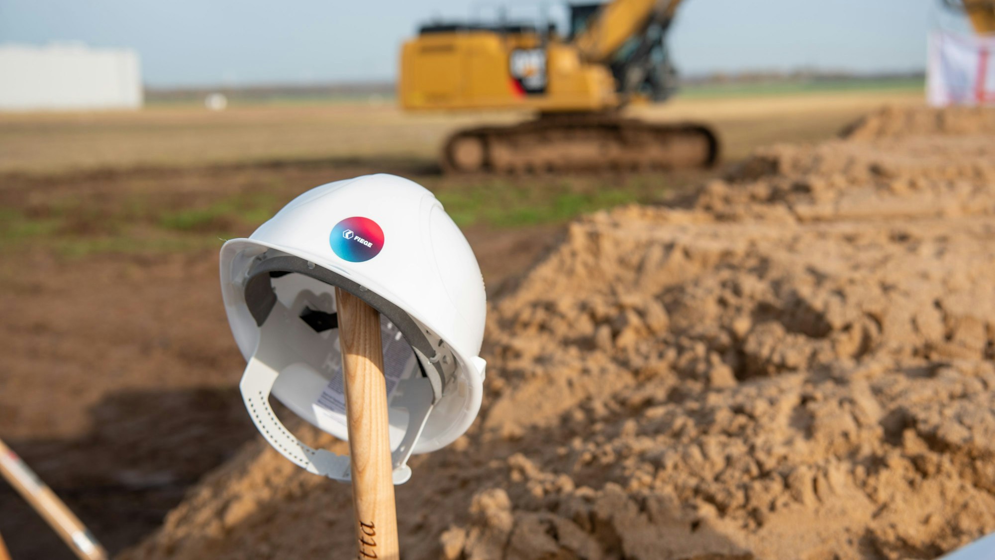 Ein Bauhelm ist auf einem Schaufelstiel abgelegt, im Hintergrund steht ein Bagger.