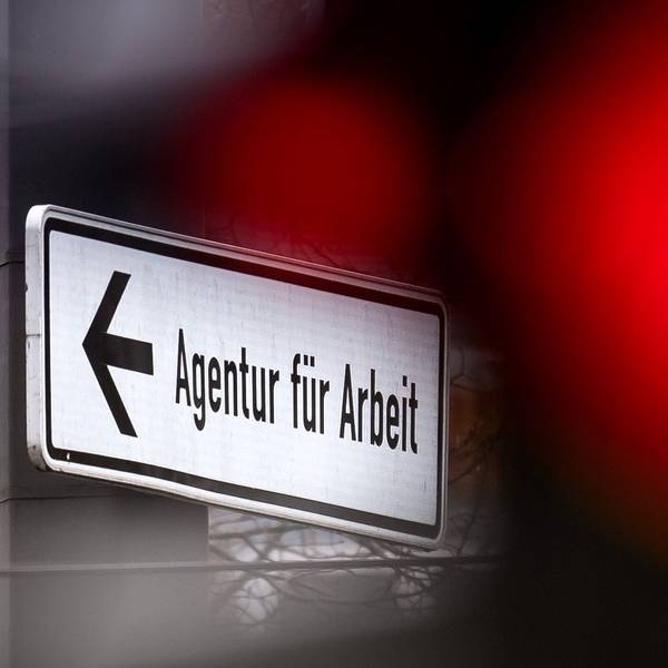 Eine rote Ampel leuchtet vor einem Schild, das auf die Agentur für Arbeit hinweist.