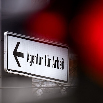 Eine rote Ampel leuchtet vor einem Schild, das auf die Agentur für Arbeit hinweist.