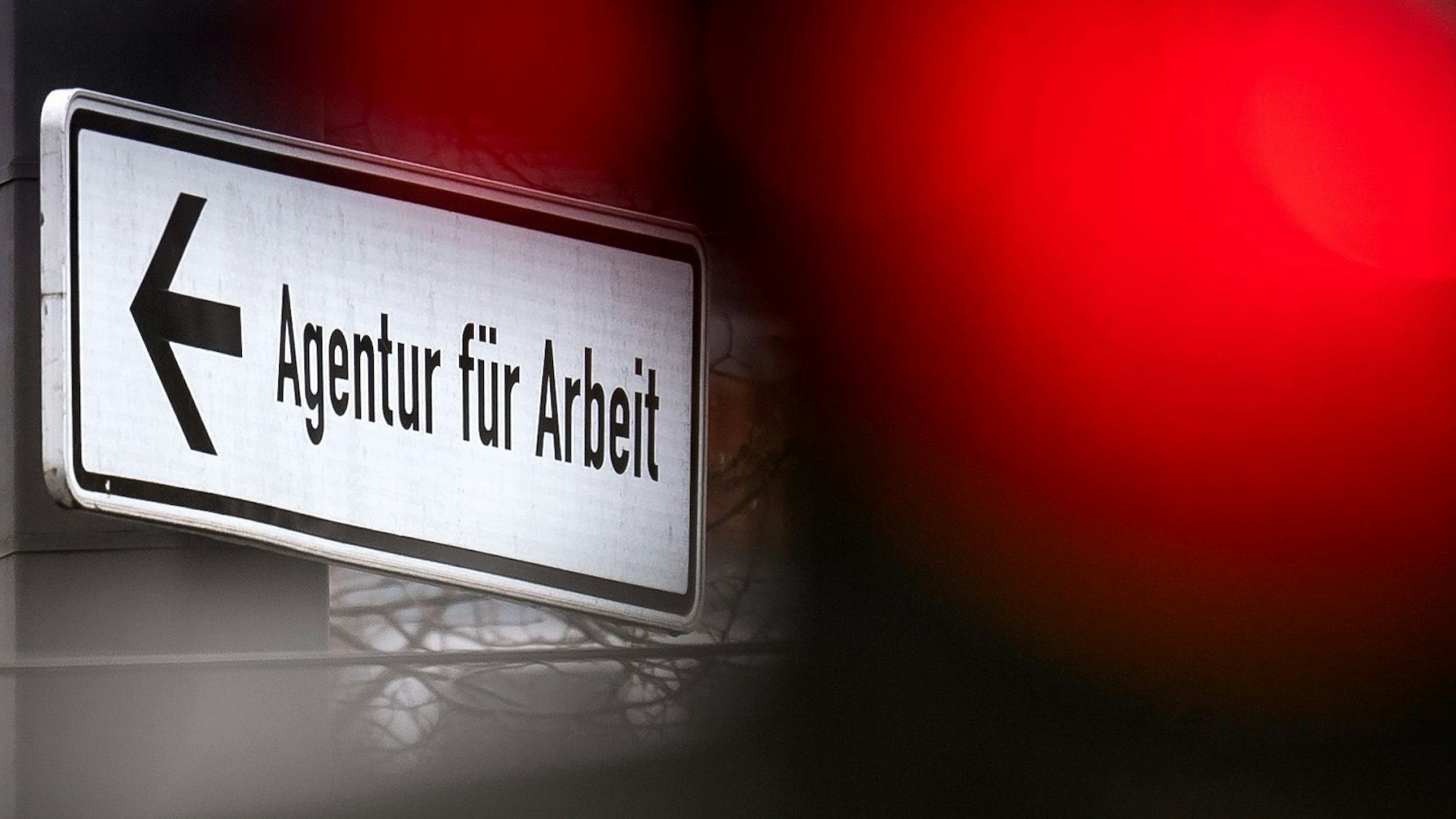 Eine rote Ampel leuchtet vor einem Schild, das auf die Agentur für Arbeit hinweist.