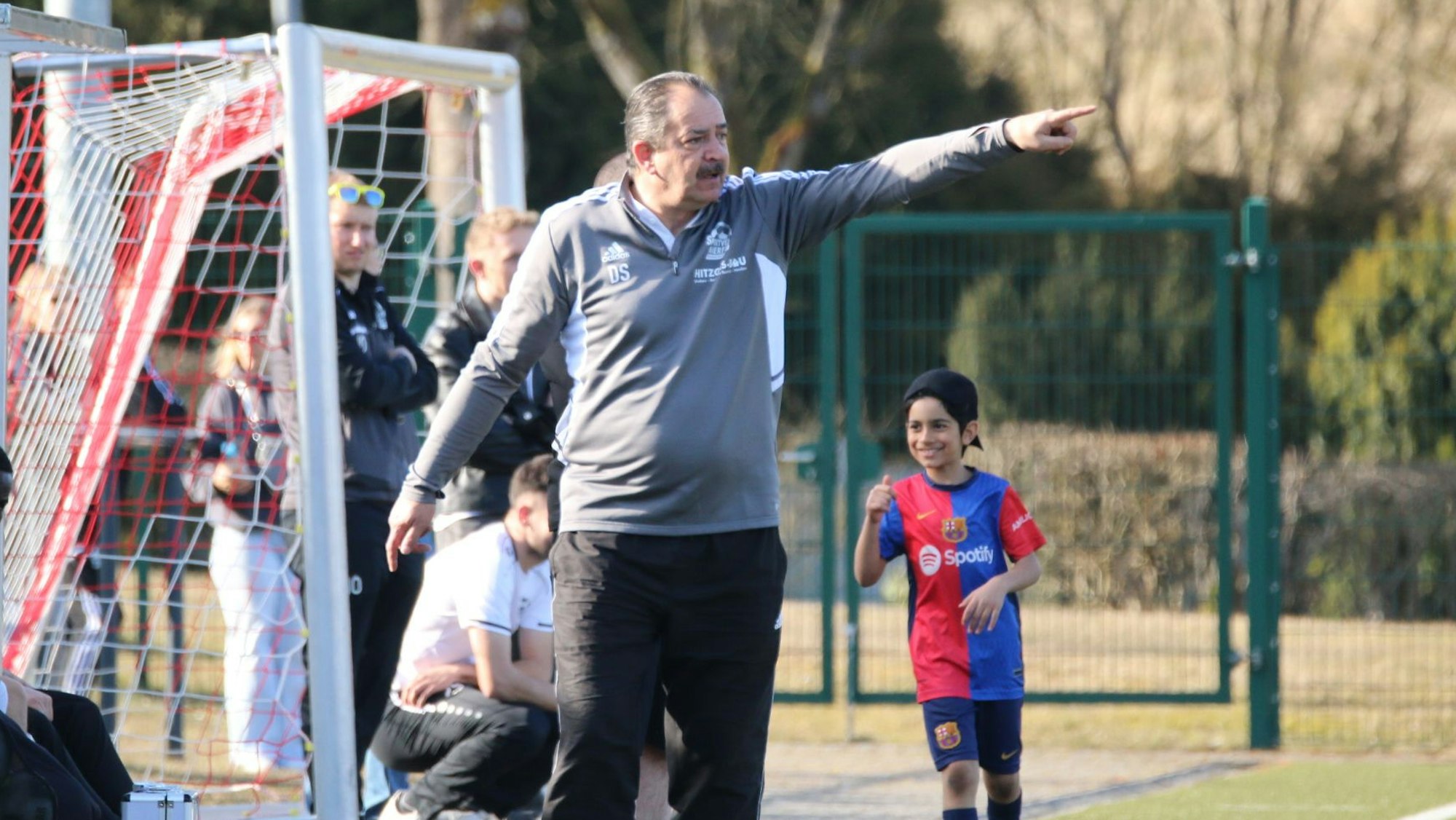Trainer Dirk Scheer deutet mit ausgestrecktem Arm aufs Spielfeld. Links sind ein Fußballtor und Zuschauer zu sehen.