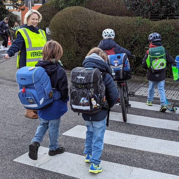 Das Foto zeigt eine Mutter, die Kindern über einen Zebrastreifen hilft.