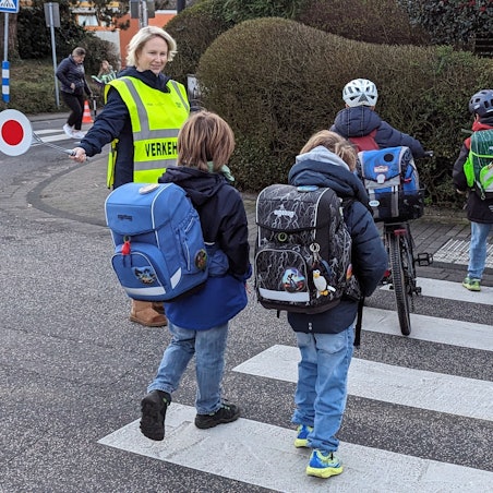Das Foto zeigt eine Mutter, die Kindern über einen Zebrastreifen hilft.