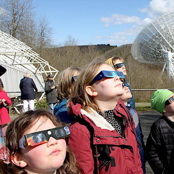 Zahlreiche Menschen tragen Schutzbrillen und betrachten die partielle Sonnenfinsternis. Im Hintergrund ist das Radioteleskop Effelsberg zu sehen.