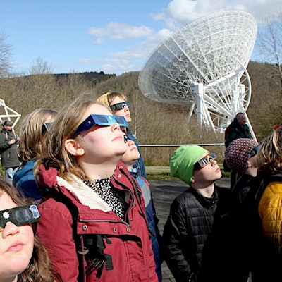 Zahlreiche Menschen tragen Schutzbrillen und betrachten die partielle Sonnenfinsternis. Im Hintergrund ist das Radioteleskop Effelsberg zu sehen.