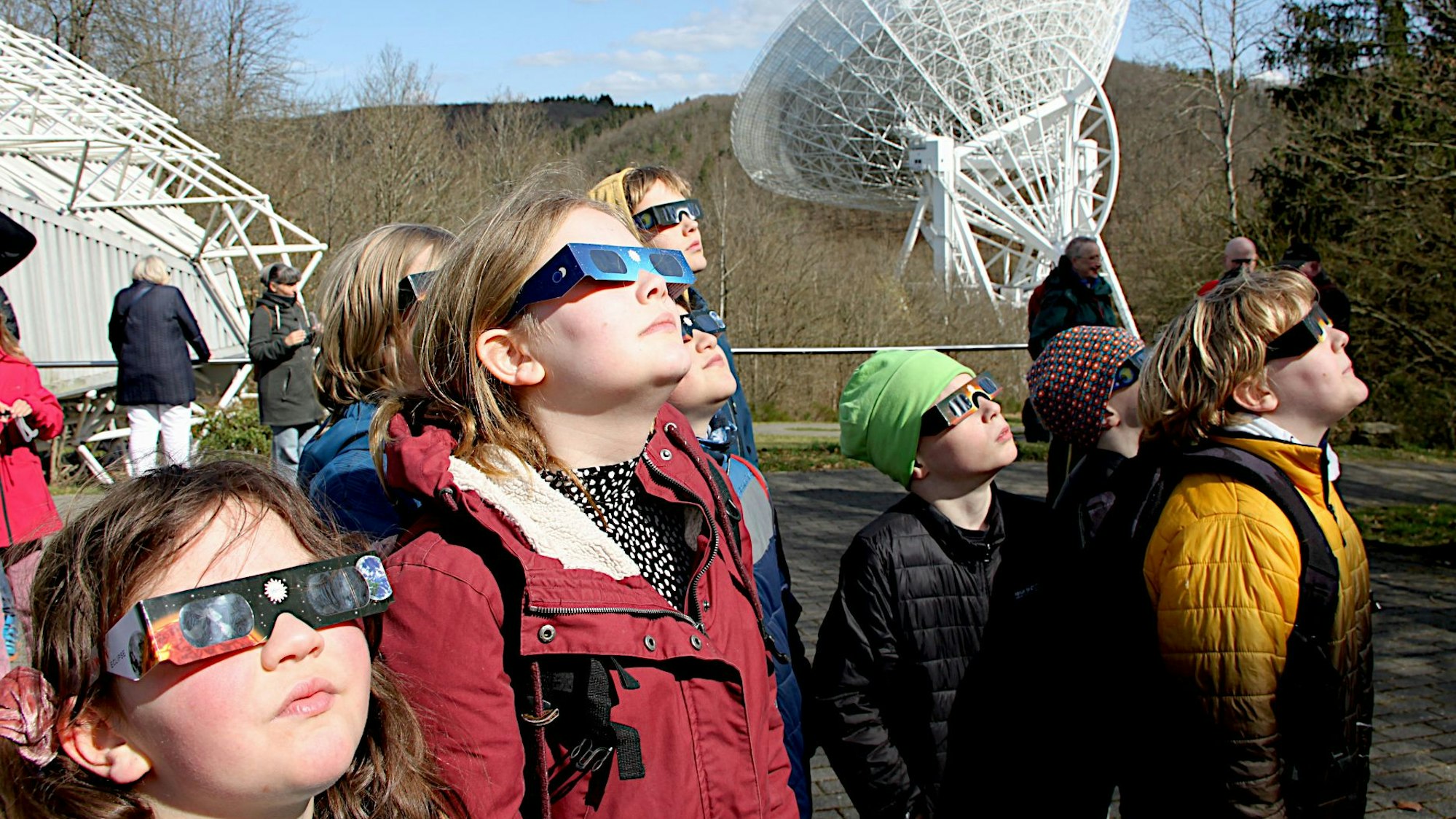 Zahlreiche Menschen tragen Schutzbrillen und betrachten die partielle Sonnenfinsternis. Im Hintergrund ist das Radioteleskop Effelsberg zu sehen.