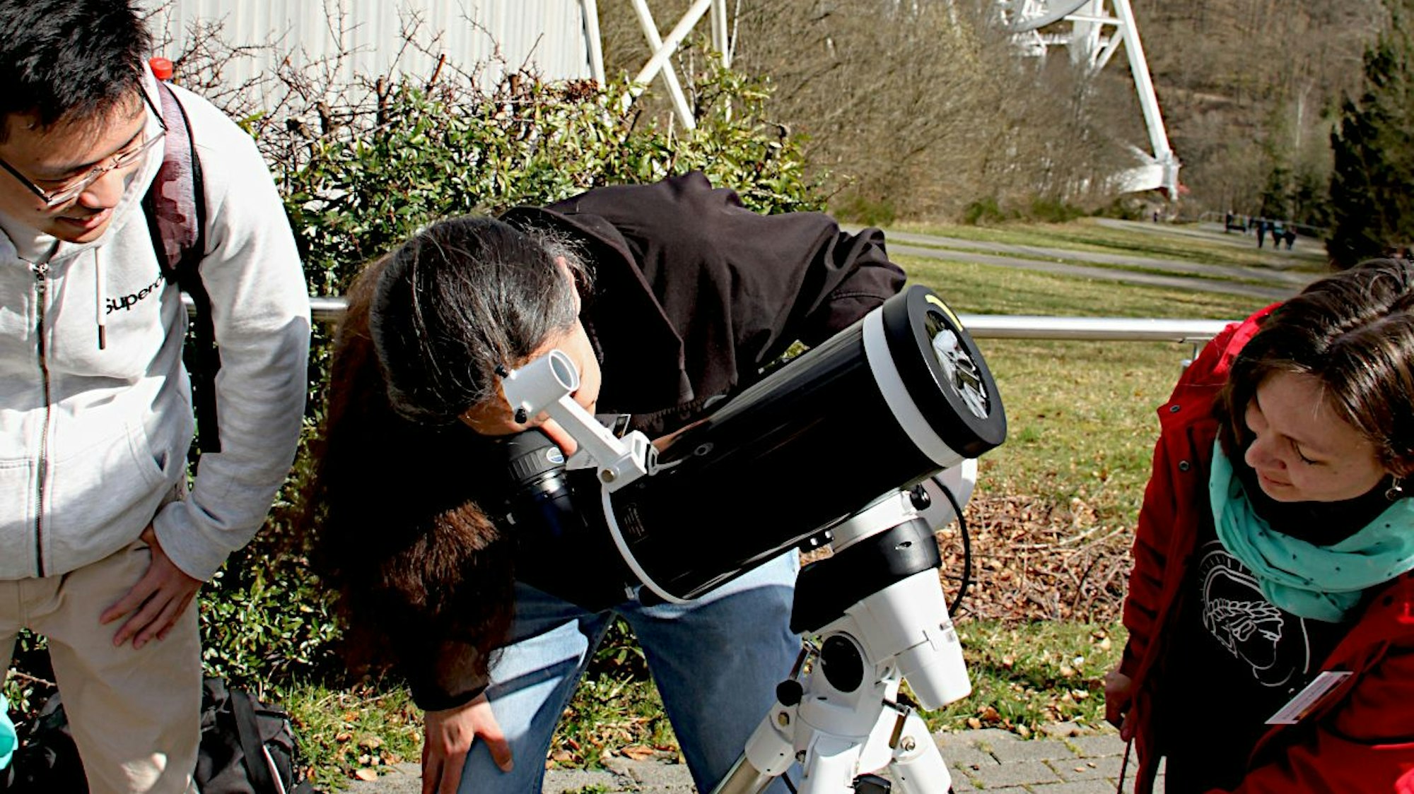 Ein Mann schaut durch ein Teleskop. Ein weiterer Mann und eine Frau stehen rechts und links von ihm und schauen dabei zu.