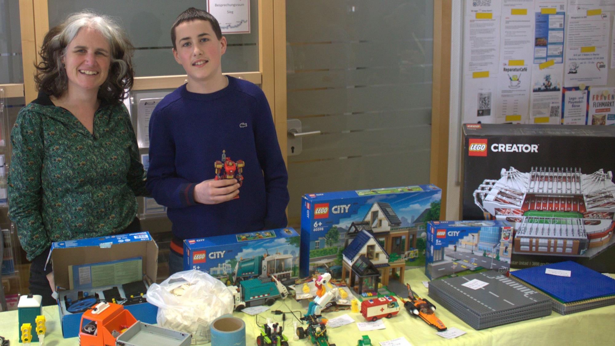 Eine Frau und ein Junge stehen hinter einem Verkaufstisch, auf dem sie Lego-Packungen und aufgebaute Modelle zeigen.