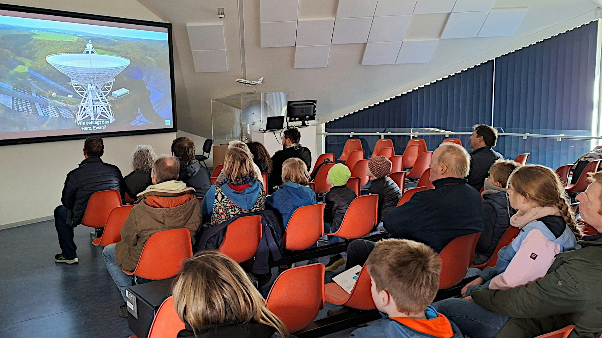 Zahlreiche Menschen sitzen in einem großen Raum auf roten Stühlen. Sie schauen auf einen Bildschirm, auf dem das Radioteleskop bei Bad Münstereifel-Effelsberg zu sehen ist.