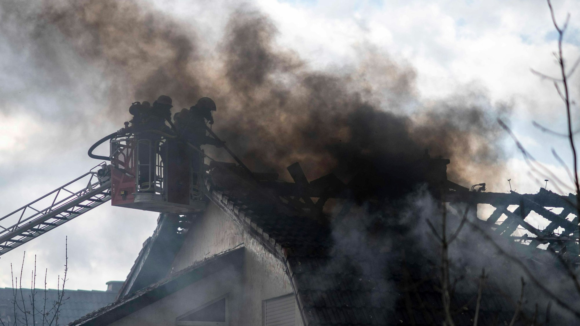 Zwei Feuerwehrleute stehen im Korb einer Drehleiter und arbeiten an einem stark qualmenden Dach.