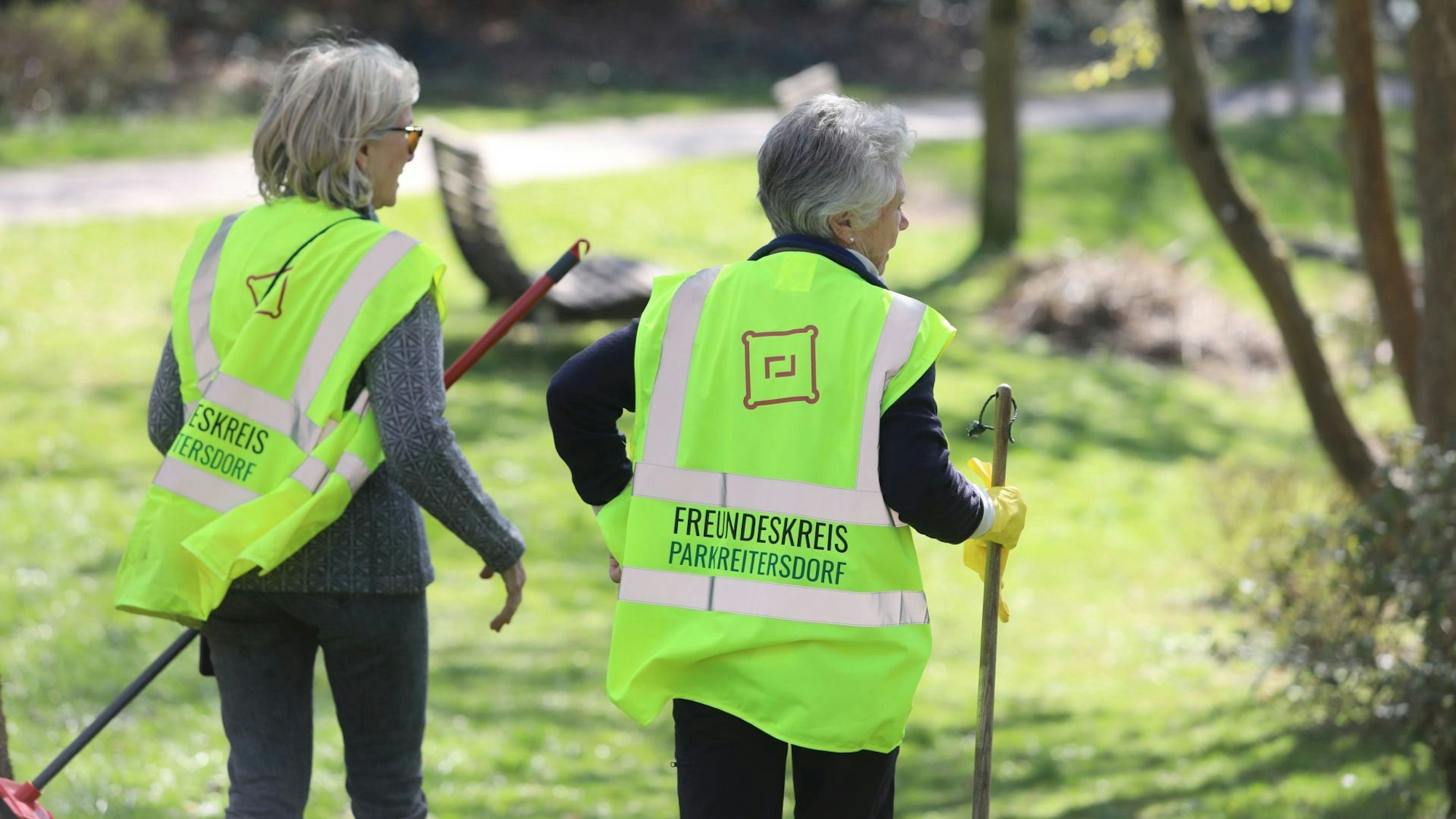 Zwei Frauen mit grauen Haaren. Sie halten Besen in der Hand und tragen reflektierende Westen mit der Aufschrift Freundeskreis Park Reitersdorf.