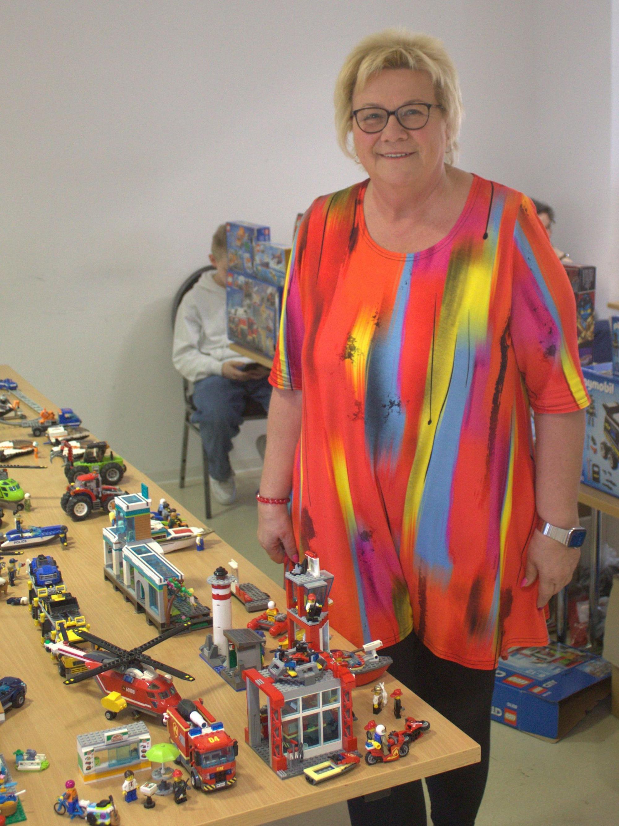 Eine blonde Frau in einer bunten Bluse steht neben einem Tisch mit Lego-Modellen.