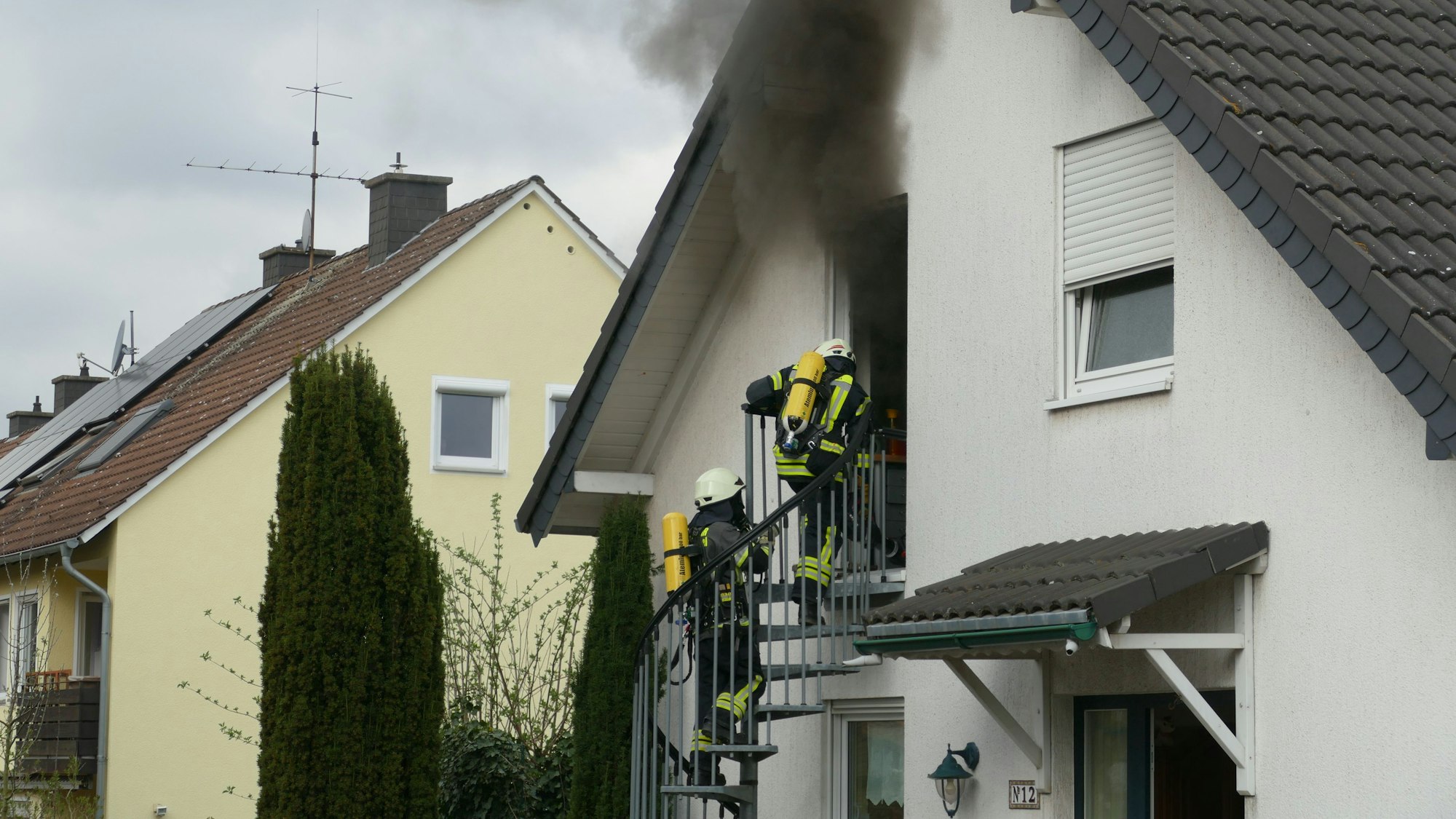 Einsatzkräfte der Feuerwehr stehen auf einer Wendeltreppe; sie sind mit Atemschutzgeräten ausgestattet, über die zwei Personen zieht dichter schwarzer Rauch.