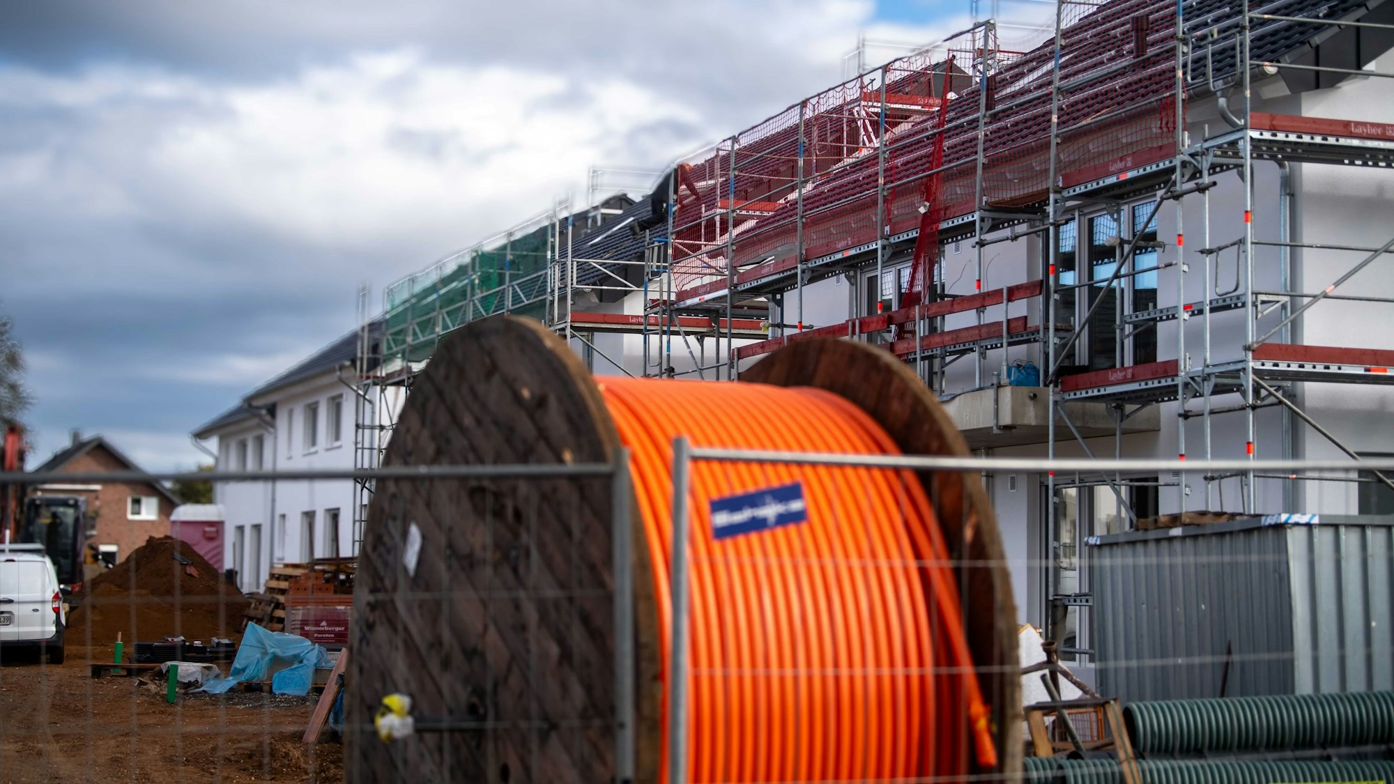 Eine große hölzerne Kabeltrommel mit einer orangefarbenen Leitung steht auf einer Baustelle. Die Neubauten im Hintergrund sind eingerüstet.
