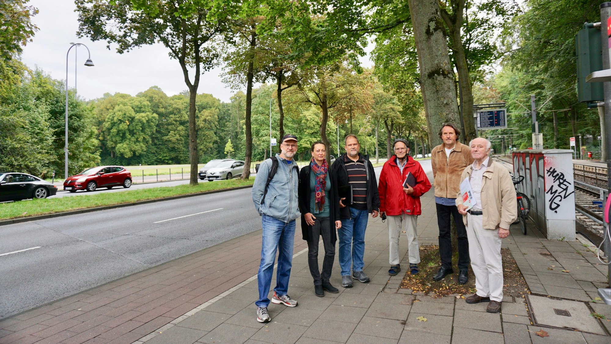 Fünf Männer und eine Frau stehen auf dem Bürgersteig an einer vierspurigen Straße, rechts von ihnen ist eine Straßenbahnhaltestelle zu sehen.