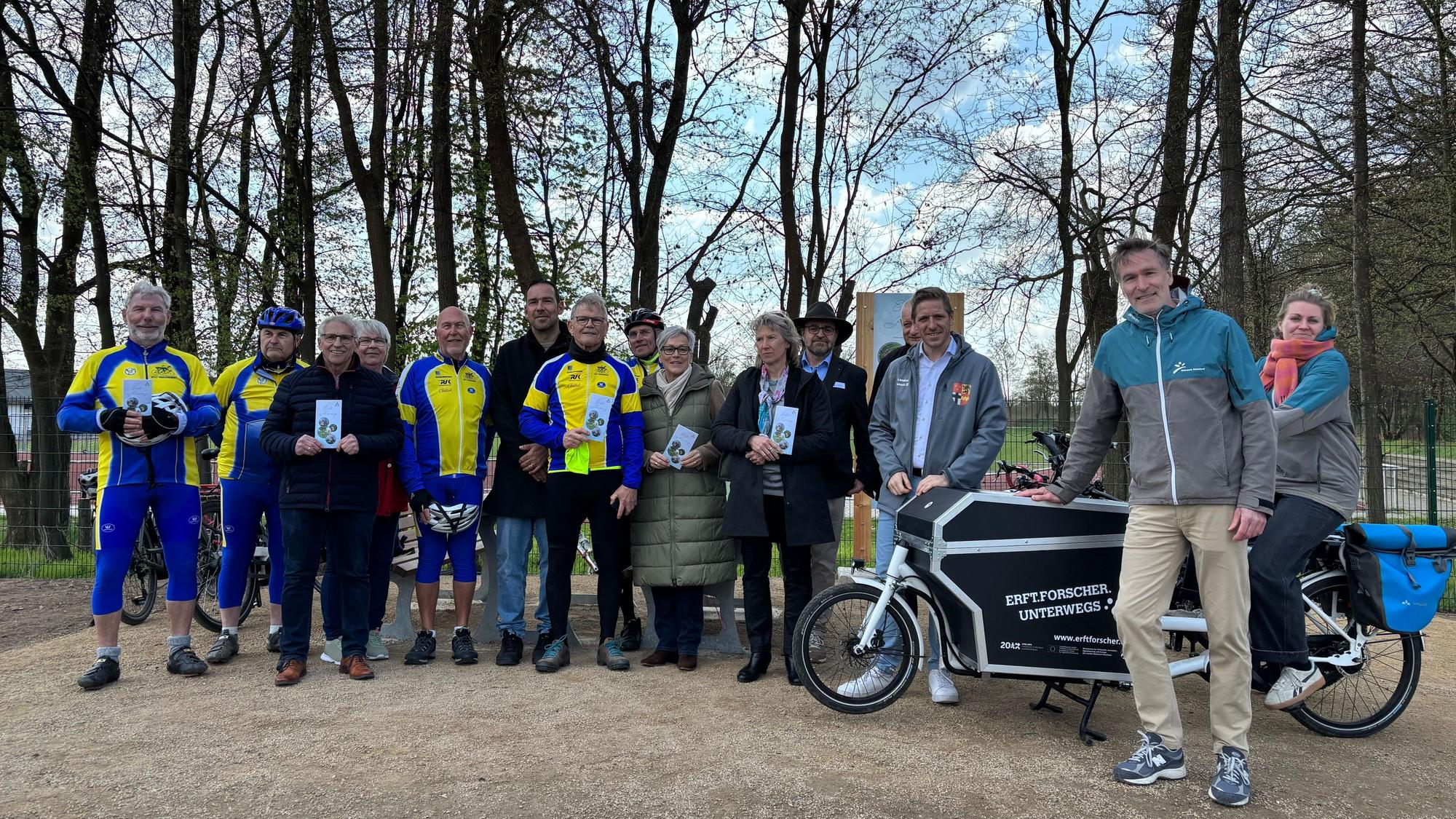Eine Gruppe, teils in Fahrradanzügen, posiert im Freien vor einem Schild.