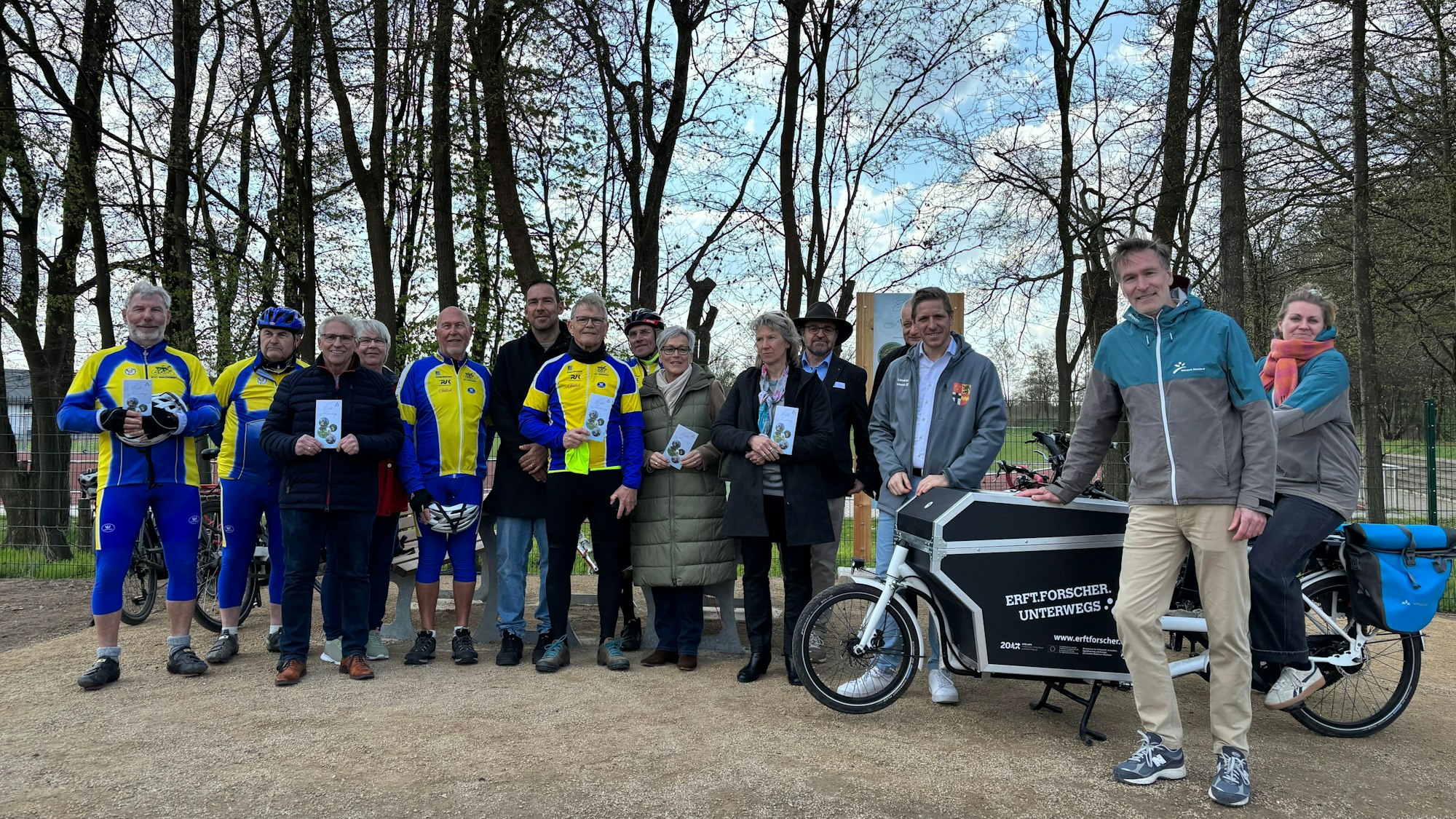 Eine Gruppe, teils in Fahrradanzügen, posiert im Freien vor einem Schild.