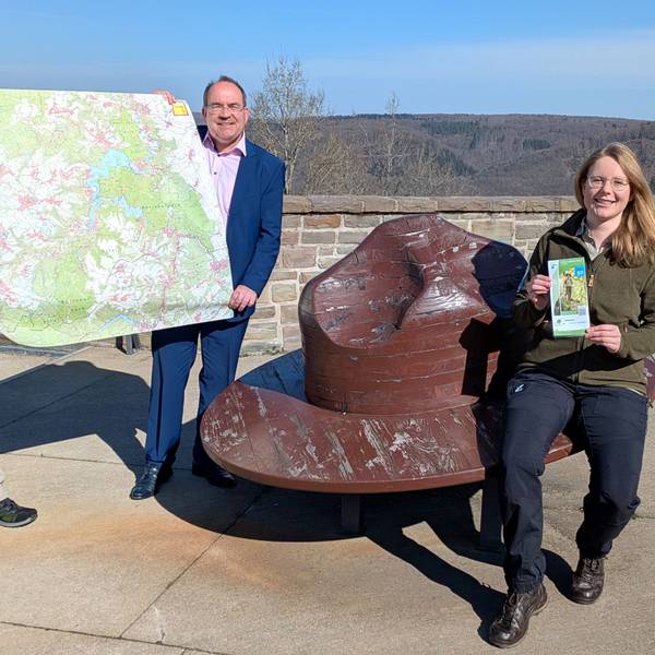 Michael Lammertz und Ralf Nolten (v.l.) präsentieren die neue Nationalpark-Wanderkarte; Jasmin Daus sitzt auf einer Holzbank, die einem Rangerhut nachempfunden ist