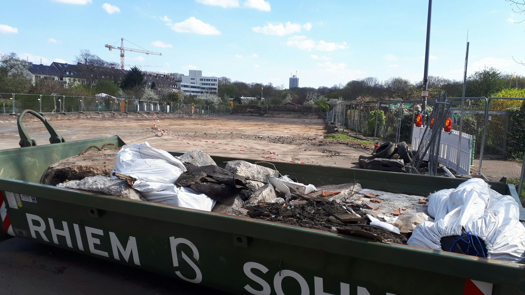 Hinter einem Container mit Bauschutt ist eine große abgesperrte freie Fläche zu sehen.