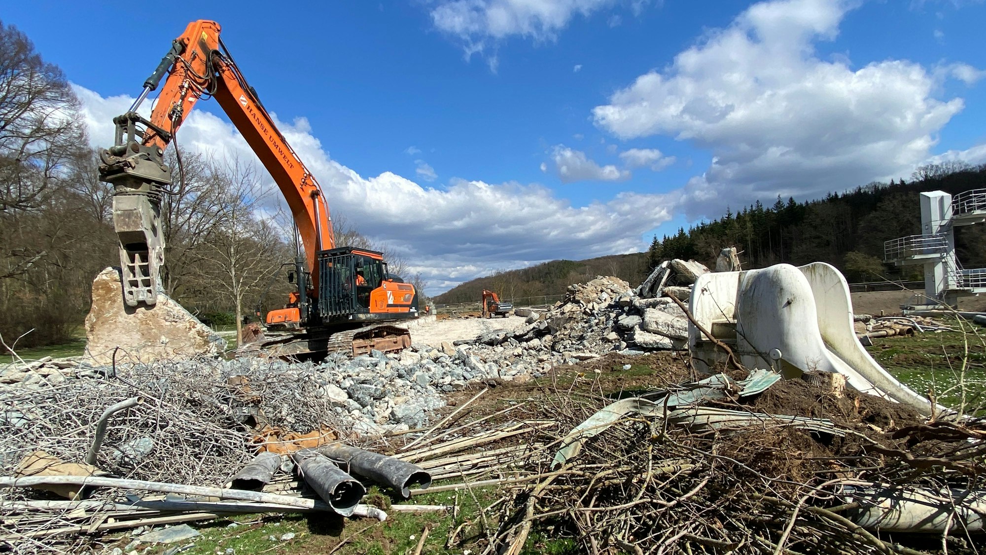Ein Bagger zerkleinert große Betonstücke. Auf dem Boden liegt Schutt und Metallschrott. Mitten drin steht eine weiße Elefantenrutsche.