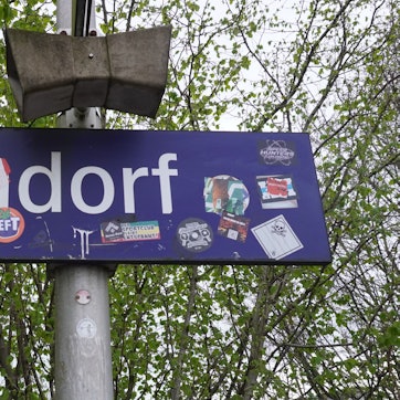 Das Foto zeigt ein Bahnhofsschild mit der Aufschrift „Sindorf“, die teils überklebt ist.