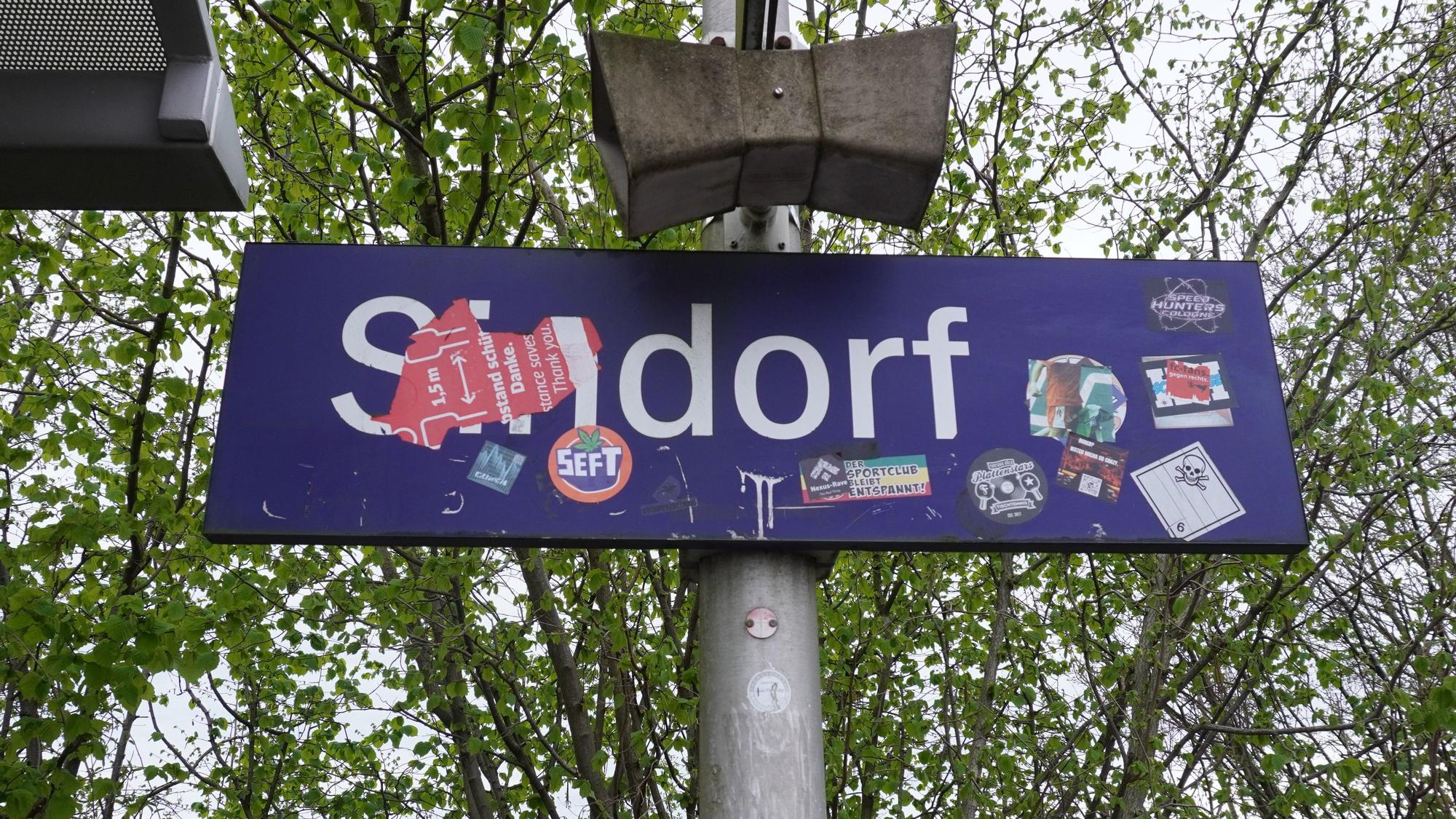 Das Foto zeigt ein Bahnhofsschild mit der Aufschrift „Sindorf“, die teils überklebt ist.
