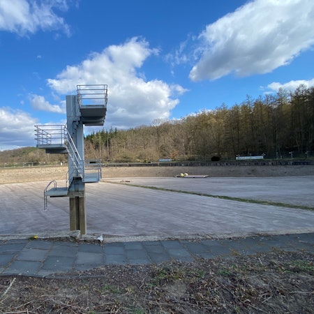 Ein leer gelaufenes, sehr tiefes Schwimmbecken, darin steht ein alter Sprungturm mit drei Ebenen.