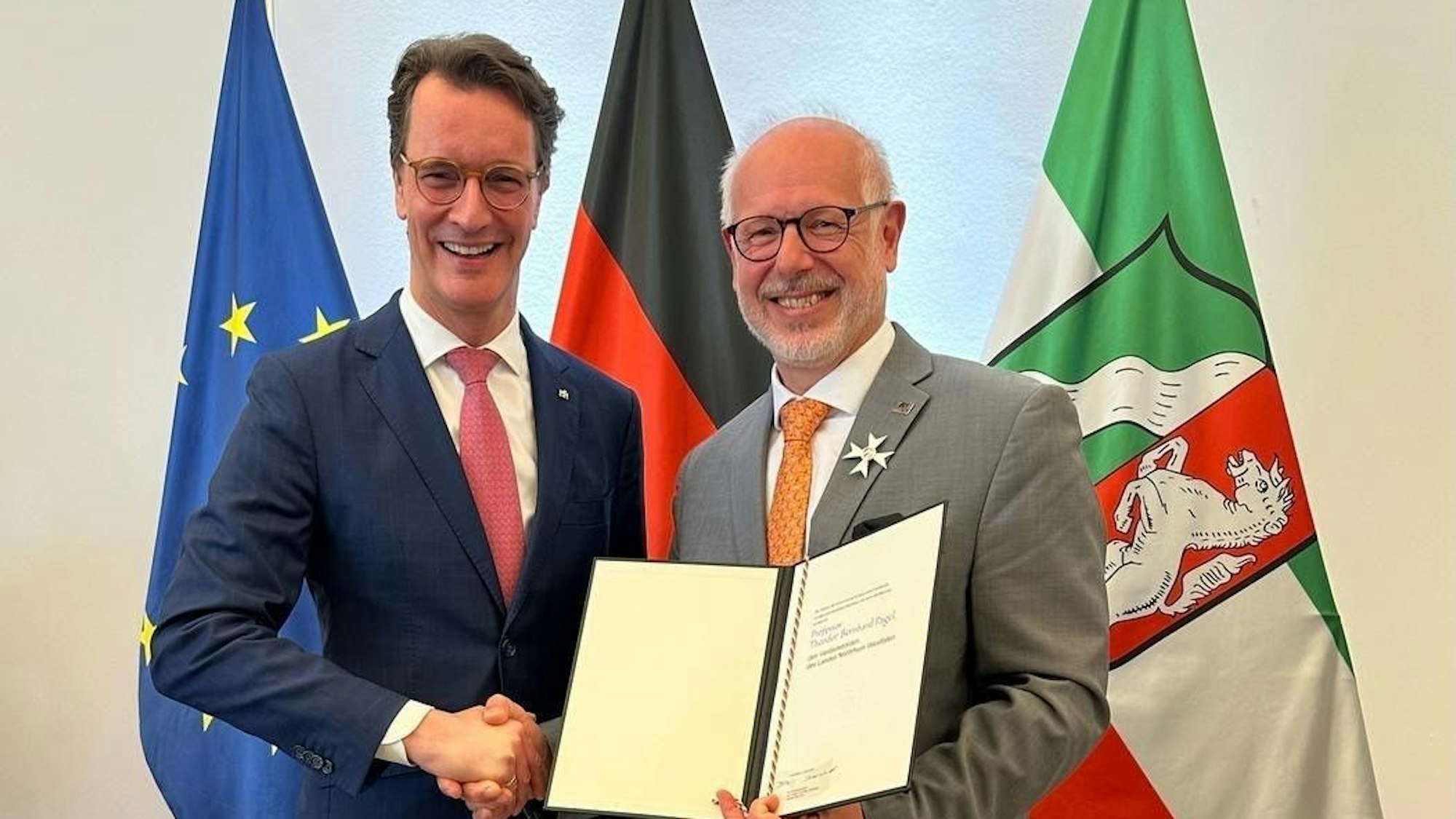 Zwei Männer mit einer Urkunde vor Flaggen.