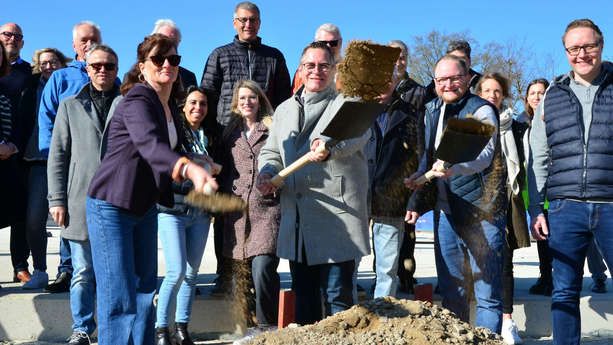 Zu sehen sind mehrere Menschen, von denen drei für den symbolischen ersten Spatenstich eine Schaufel in die Hand genommen haben.