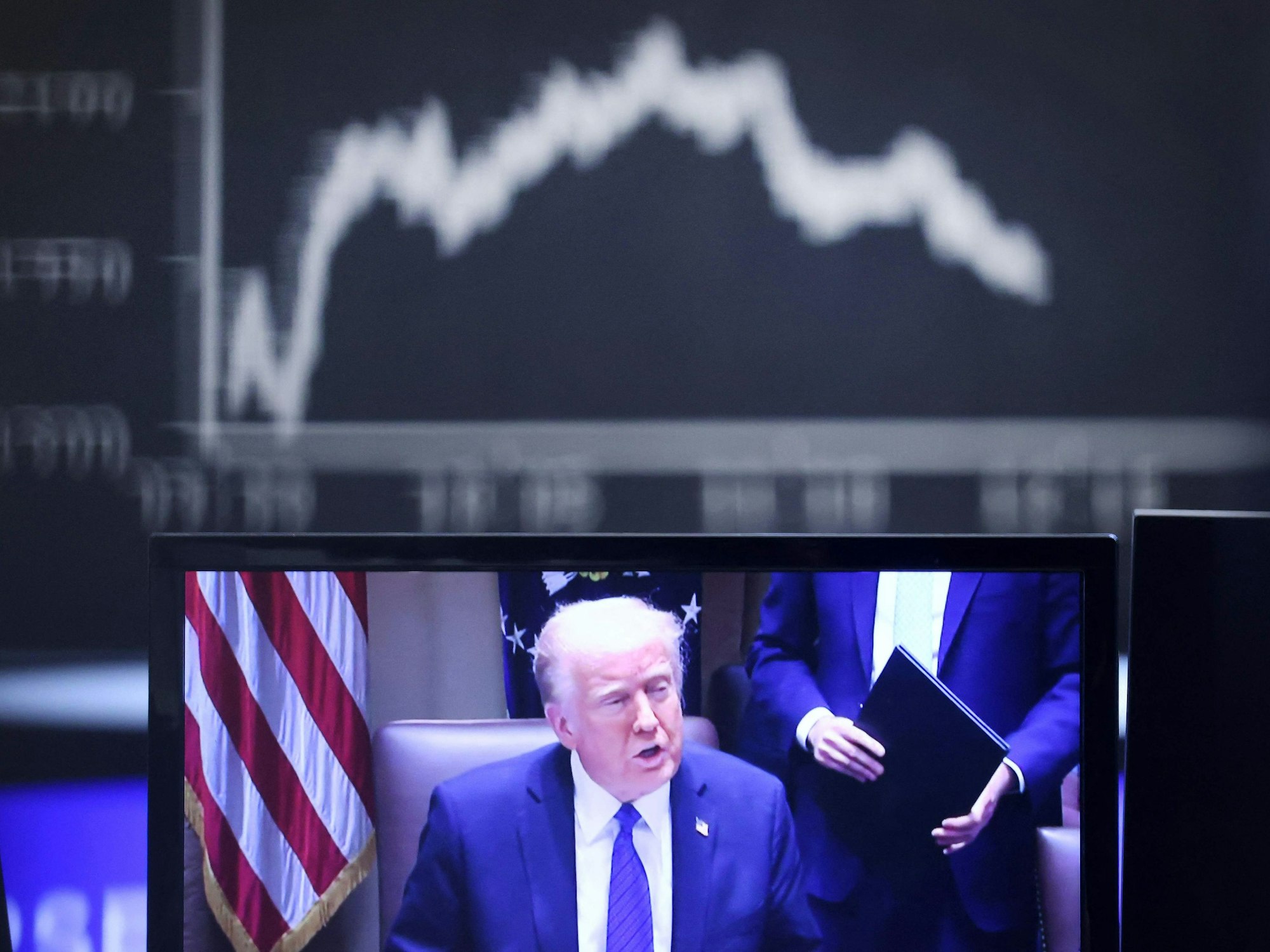 US President Donald Trump is seen on a TV screen below a display showing the German Stock Market Index DAX at the stock Donald Trumps Politik wird die US-Bürger belasten.in Frankfurt, Germany, on April 3, 2025. (Photo by Daniel ROLAND / AFP)