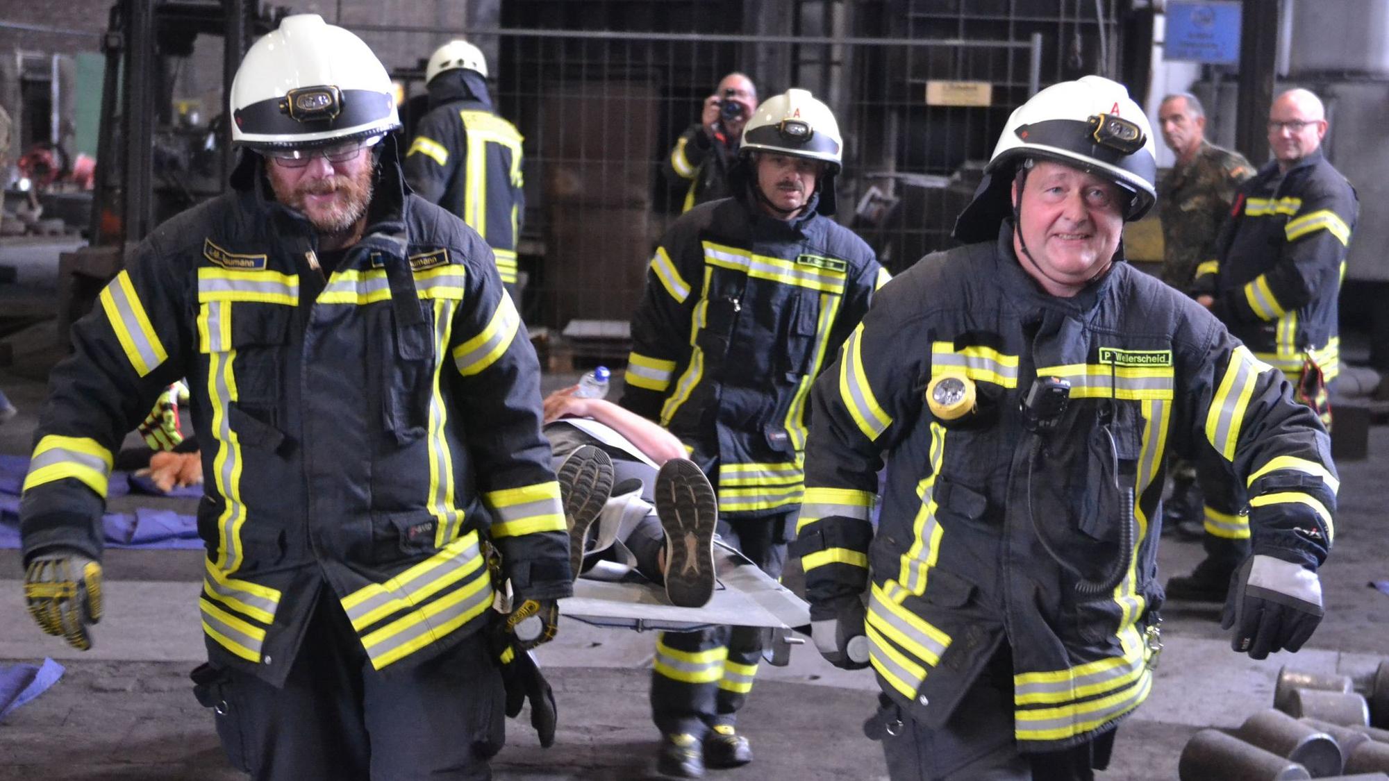 Im Rahmen einer Übung tragen vier Feuerwehrleute einen Verletztendarsteller auf einer grauen Krankentrage.