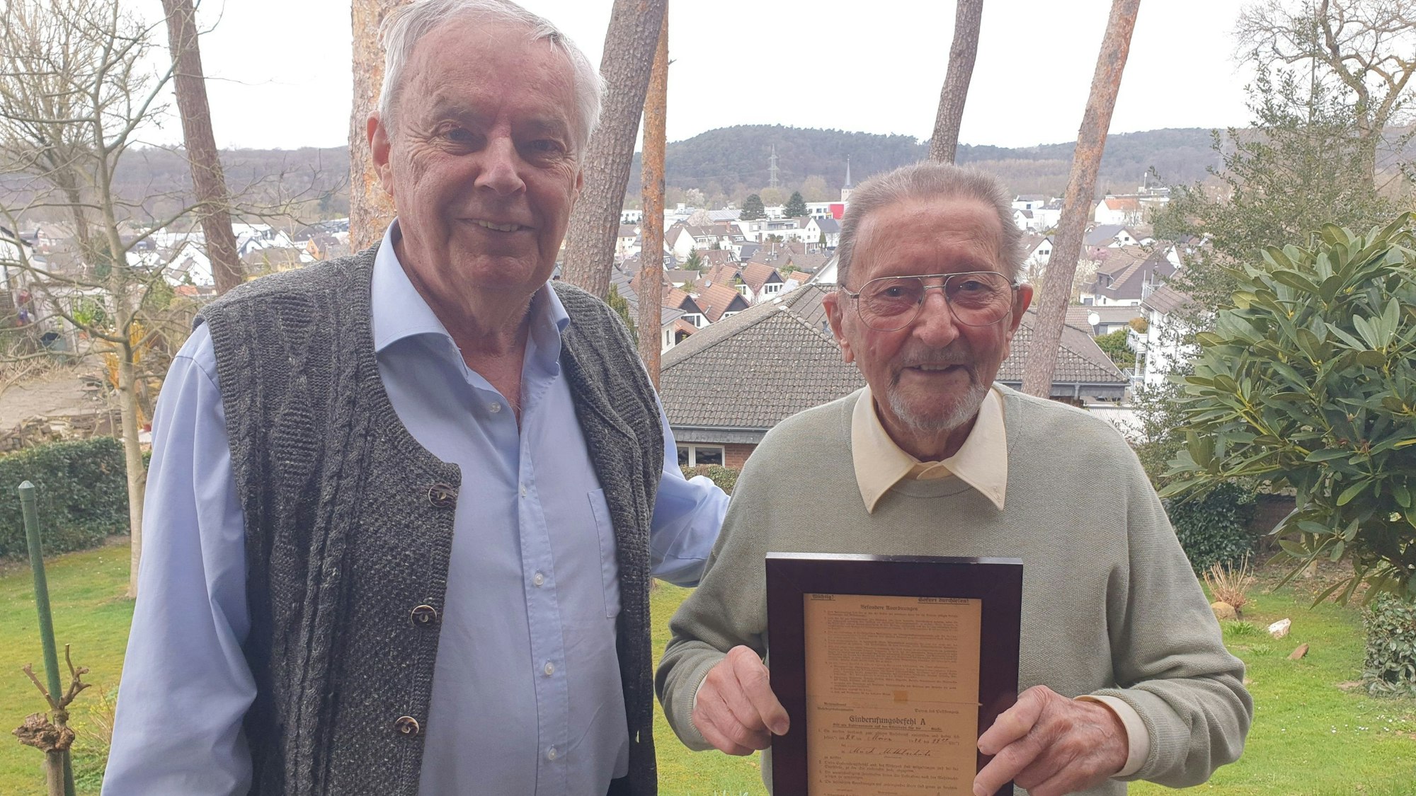Zwei betagte Männer stehen auf einer Anhöhe über einer Stadt. Der Ältere hält ein gerahmtes Dokument vor sich.