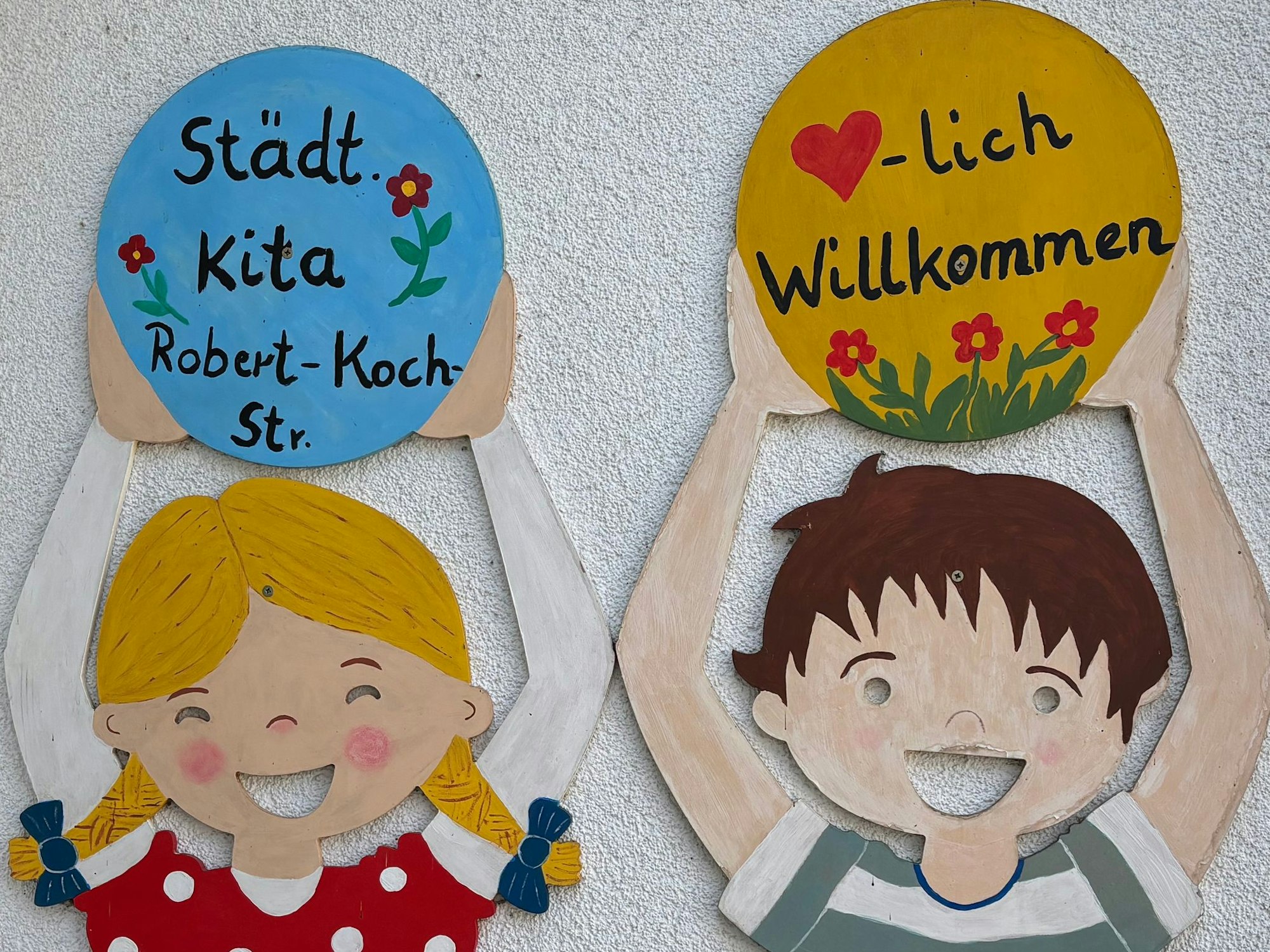 Pappaufsteller eines Mädchens mit Zöpfen und eines Jungen halten Schilder in den Händen, auf denen „Städt. Kita Robert-Koch-Str.“ und „Herzlich Willkommen“ steht.