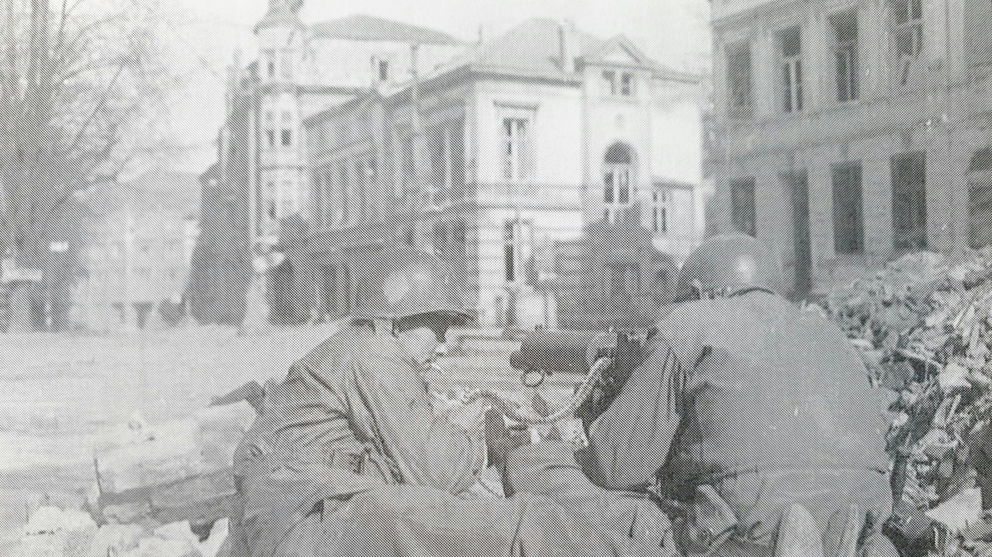 Ein Schwarz-Weiß-Foto von Soldaten in einer Straße mit zerstörten Gebäuden, sie bedienen ein Maschinengewehr.