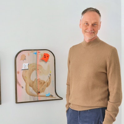Ein Mann in einem braunen Rollkragenpullover steht neben zwei Bildern. Die Werke sind vollgepackt mit unterschiedlichen Details.