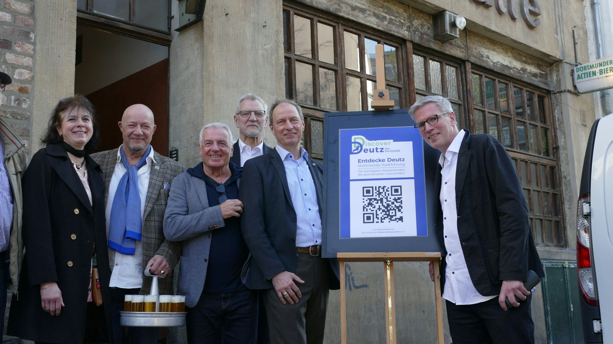 Menschen stehen vor einer Gaststätte, in ihrer Mitte ein Schild mit einem QR-Code.