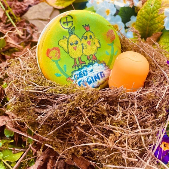 Eine gelbe Hülse der bekannten Überraschungseier liegt in einem Nest. Daneben liegt ein bunt bemalter Stein.