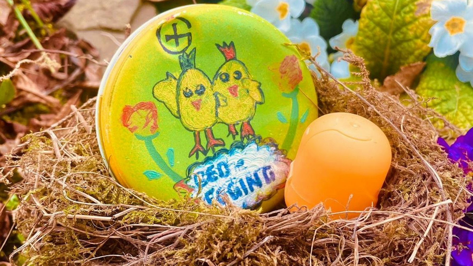 Eine gelbe Hülse der bekannten Überraschungseier liegt in einem Nest. Daneben liegt ein bunt bemalter Stein.