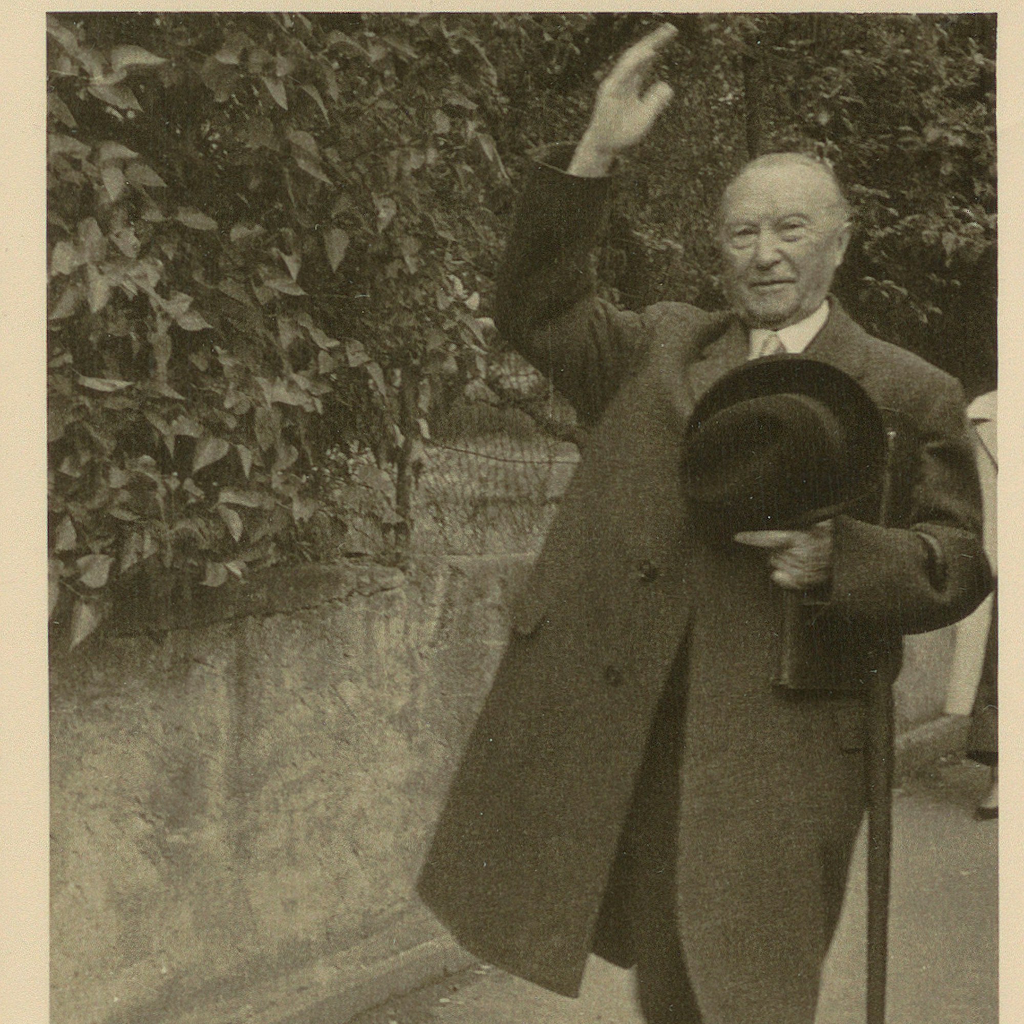 Ein altes Foto zeigt einen Mann im Mantel, der freundlich winkt und Hut, Aktentasche und Regenschirm trägt.