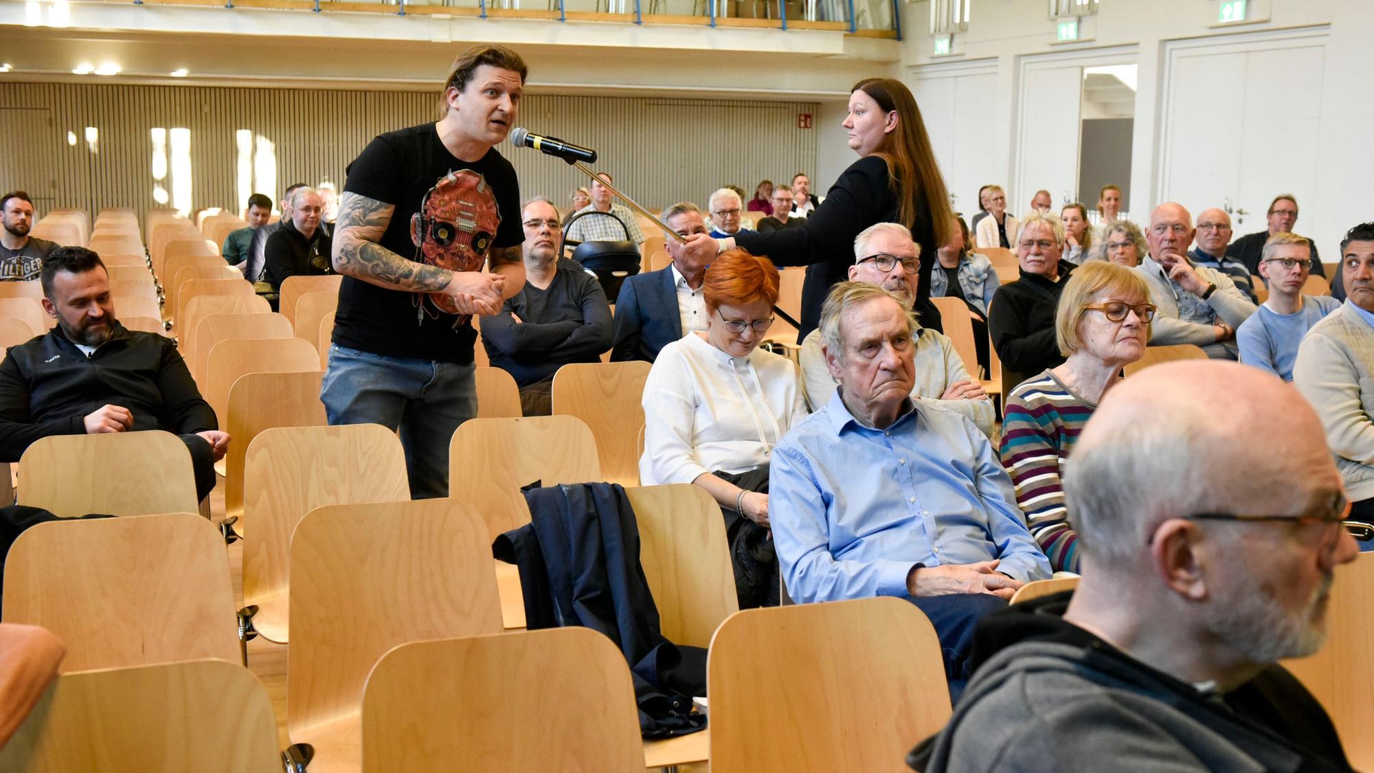 Mehrere Menschen sitzen in Stuhlreihen, ein Mann spricht in ein Mikrofon, das ihm eine Frau hinhält.