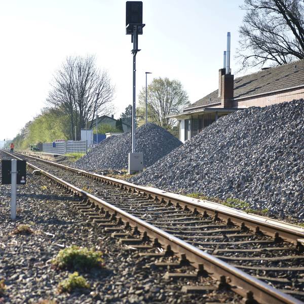 Gleisschotter liegt in großen Haufen neben den Schienen am Bahnhof Zülpich. Im Hintergrund arbeiten ein paar Bauarbeiter auf den Gleisen.