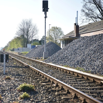 Gleisschotter liegt in großen Haufen neben den Schienen am Bahnhof Zülpich. Im Hintergrund arbeiten ein paar Bauarbeiter auf den Gleisen.