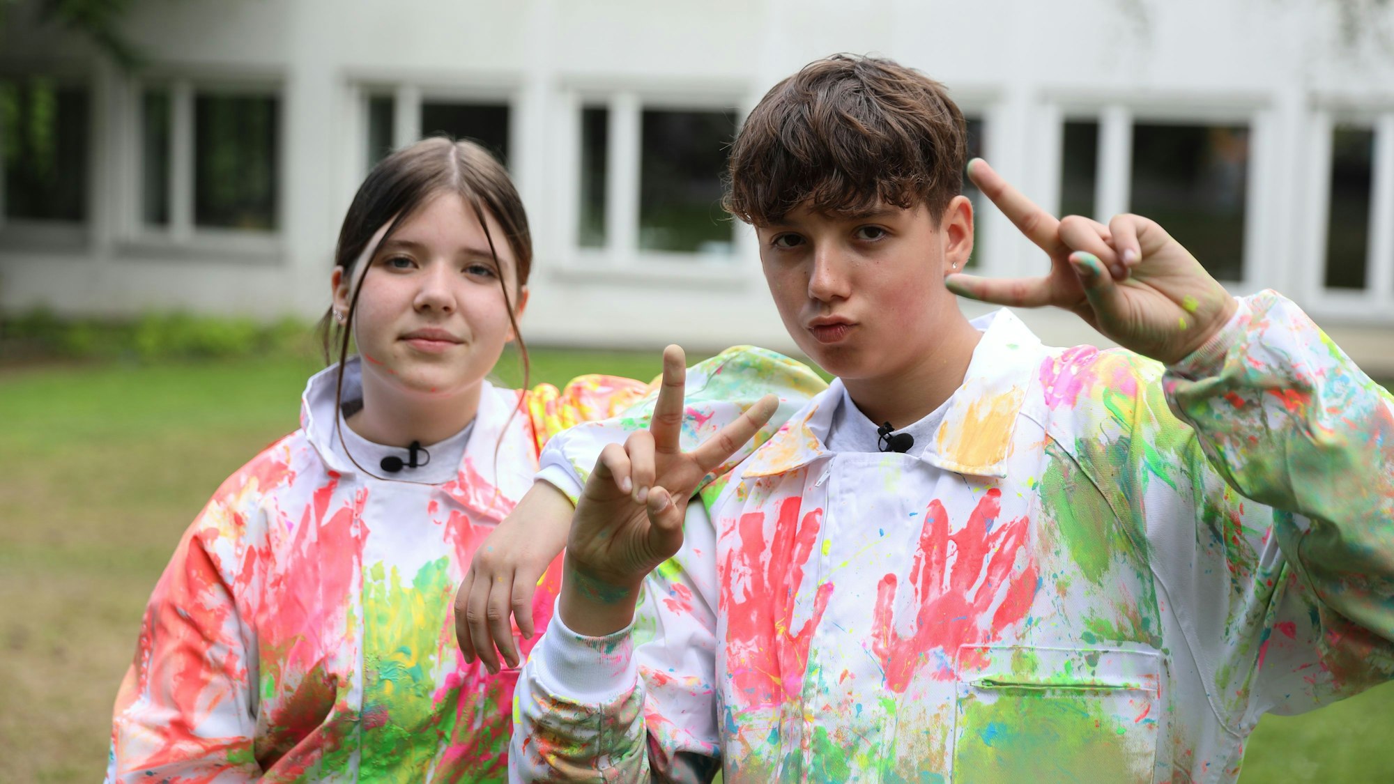 Ein Junge und ein Mädchen stehen in bunt bemalten Maleranzügen auf einer Wiese. Der Junge zeigt zwei Peace-Zeichen.