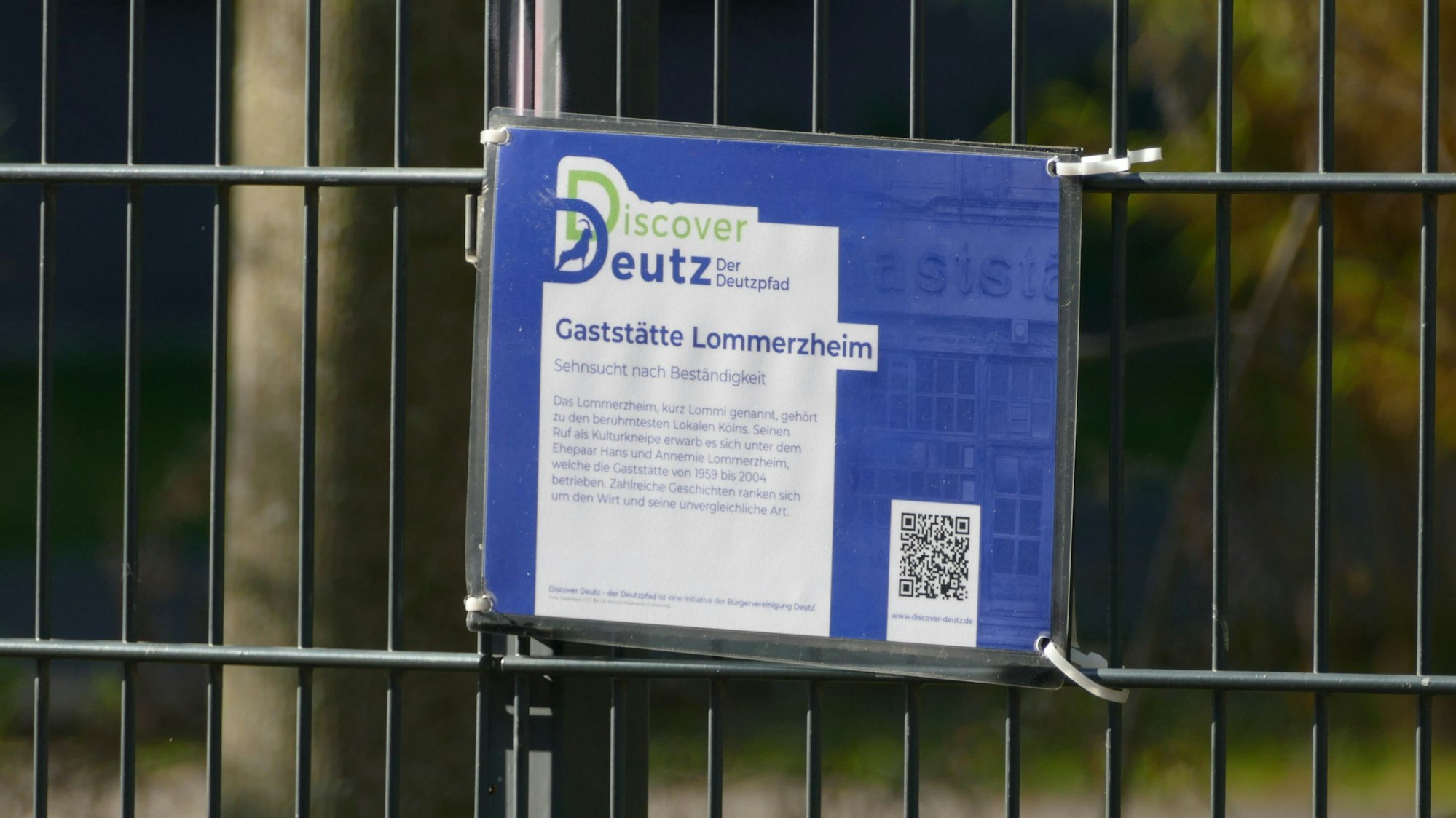 Eine Tafel mit einem QR-Code hängt an einem Zaun.