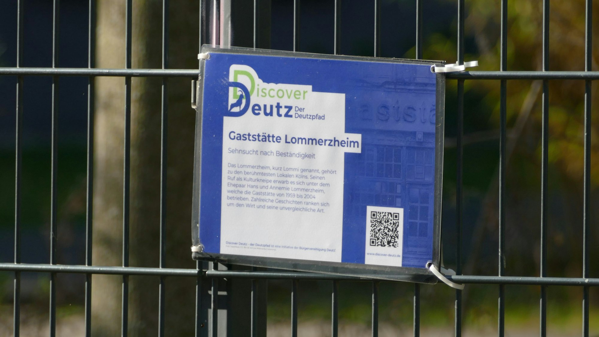 Eine Tafel mit einem QR-Code hängt an einem Zaun.