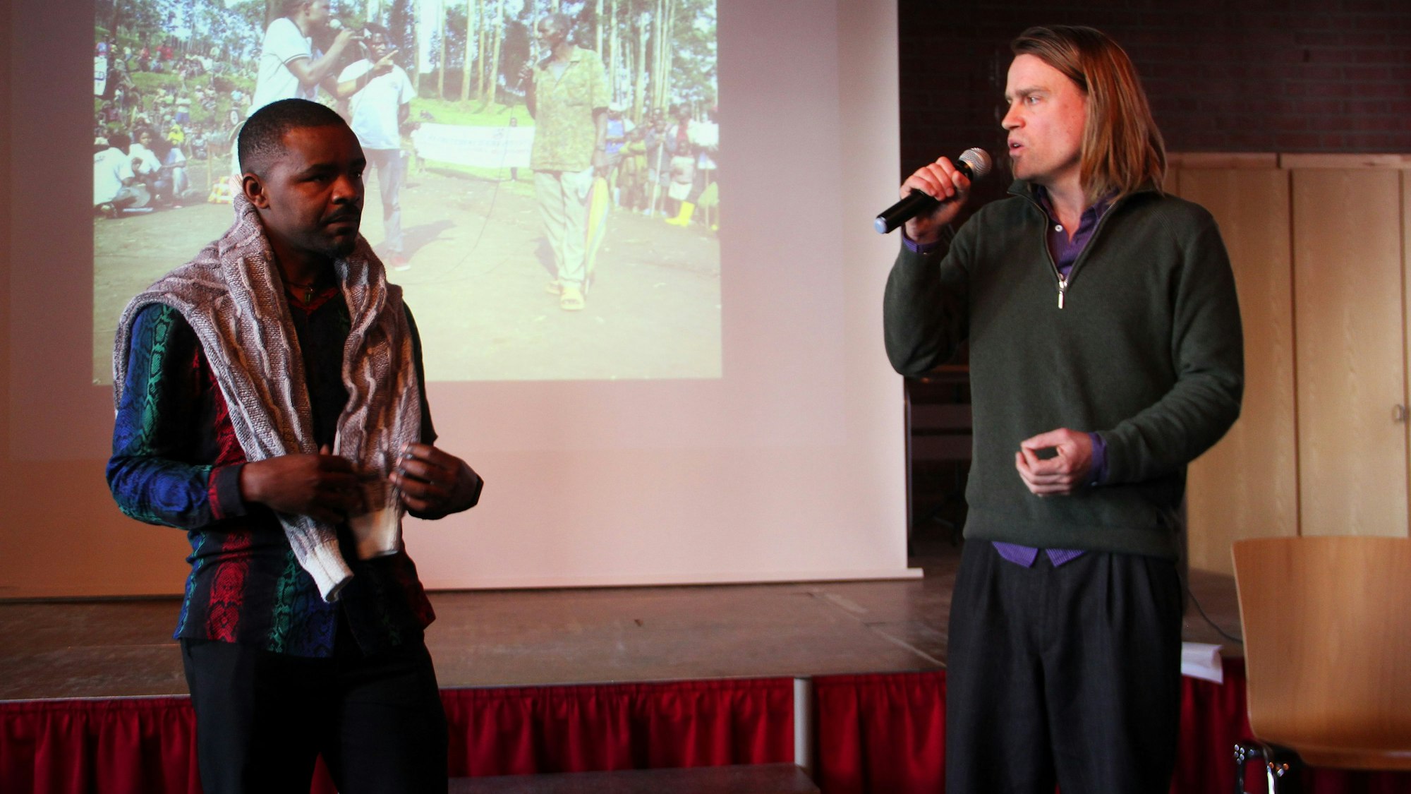 Ein dunkelhäutiger Mann und ein Mann mit schulterlangem Haar und Mikrofon in der Hand stehen vor einer Leinwand.