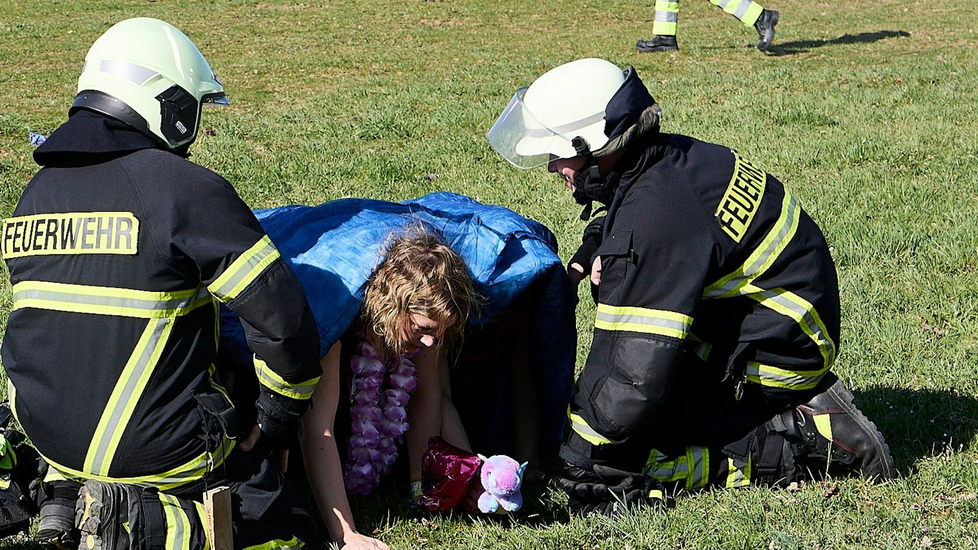 Zwei Feuerwehrleute knien im Rahmen einer Übung bei Verletztendarstellern, die sich eine Decke über den Kopf gezogen haben und über eine Wiese krabbeln.